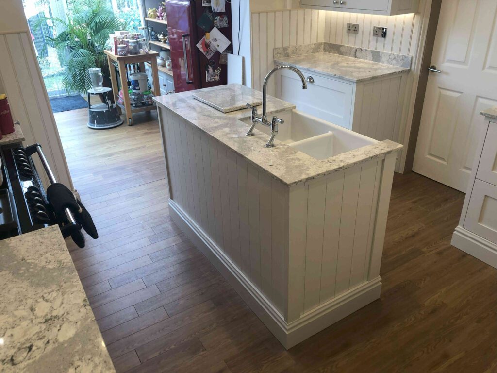 picture of a kitchen island set amongst a section of the overall kitchen. Built with a sink and with a white and grey flecked marble worktop